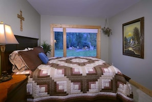 A bedroom with a large bed, a cross on the wall, a painting, and a view of the outdoors.