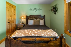 A bedroom with a wooden bed, a nightstand with a lamp, a decorative wall piece, and a potted plant.