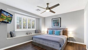 Une chambre à coucher comprenant un lit, un ventilateur de plafond, une fenêtre avec des stores, un téléviseur fixé au mur et une lampe de chevet.