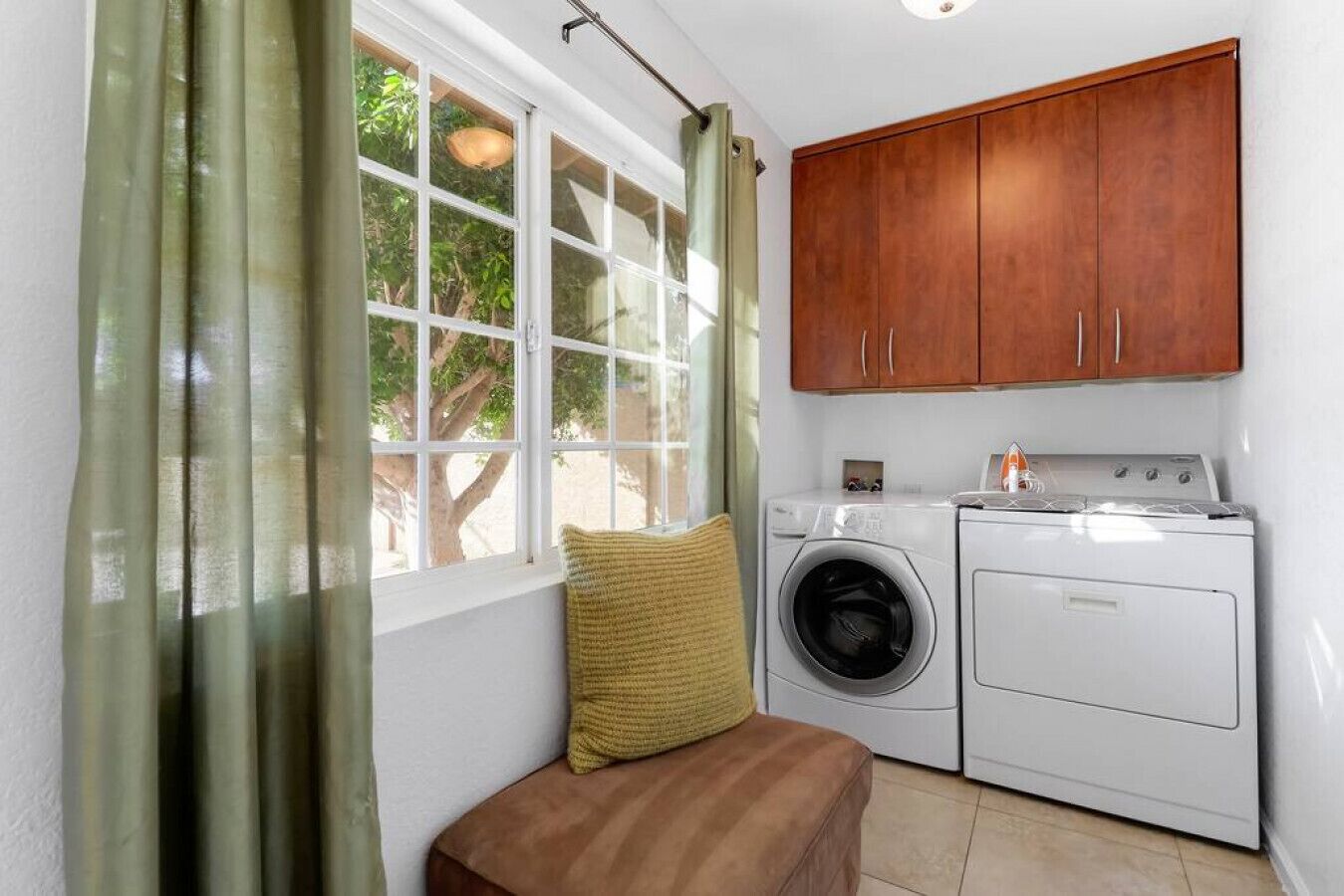 A laundry room with a washing machine and dryer, a wooden cabinet above, a window with curtains, and a bench with a cushion.
