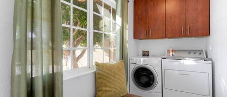 A laundry room with a washing machine and dryer, a wooden cabinet above, a window with curtains, and a bench with a cushion.
