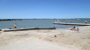 A beach with a concrete walkway leading to a pier, sandy areas, and a clear blue sky.