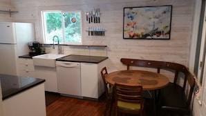 A compact kitchen with white cabinetry, a dishwasher, a sink, and a window with a view of greenery. There is a set of knives hanging on the wall and a painting above the dining table. - Your Home away from Home (Harbert)