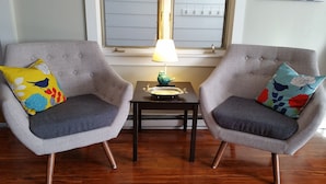 Two grey armchairs with floral cushions, a small table with a lamp, and a window with blinds. - Your Home away from Home (Harbert)