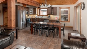 A spacious living area with a high wooden ceiling, a central dining table surrounded by chairs, and a kitchen in the background. - Cranberry Cabin Rental (Valemount)