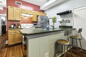 A kitchen with wooden cabinets, a stainless steel refrigerator, a microwave, and a sink with a faucet. There are two bar stools and a countertop with a green backsplash.