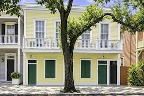 A two-story yellow building with green shutters, a white balcony, and a tree in front.