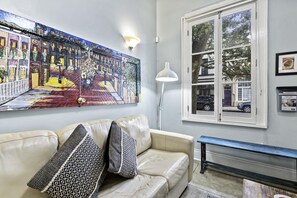 A living room with a beige sofa, patterned cushions, a large colorful painting, a white floor lamp, a window with a view of the street, and a framed picture on the wall.