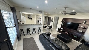 A modern living room with a black leather sofa, a flat-screen TV, and a dining area with a round table and bar stools.