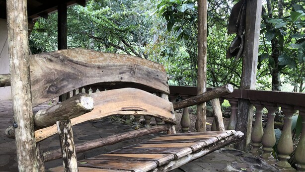 A rustic wooden bench with a curved backrest and a slatted seat, situated on a stone-paved area under a wooden structure with a railing. - Rancho el Rio: Nature, Riverside & Farm (Monteverde)