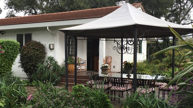 A covered patio with a table and chairs, surrounded by lush greenery and a white building with a tiled roof. - Private Cottage, Amazing Gardens within a Gated Estate  - True European Charm! (Soquel)