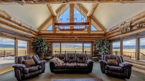 L’intérieur d’une cabane en rondins est aménagé avec un grand canapé en cuir, deux fauteuils et une table basse. De grandes fenêtres offrent une vue imprenable sur les montagnes.