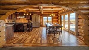 L’intérieur d’une cabane en rondins, avec un plancher en bois, une cuisine, un coin repas et de grandes fenêtres offrant une vue sur les montagnes.