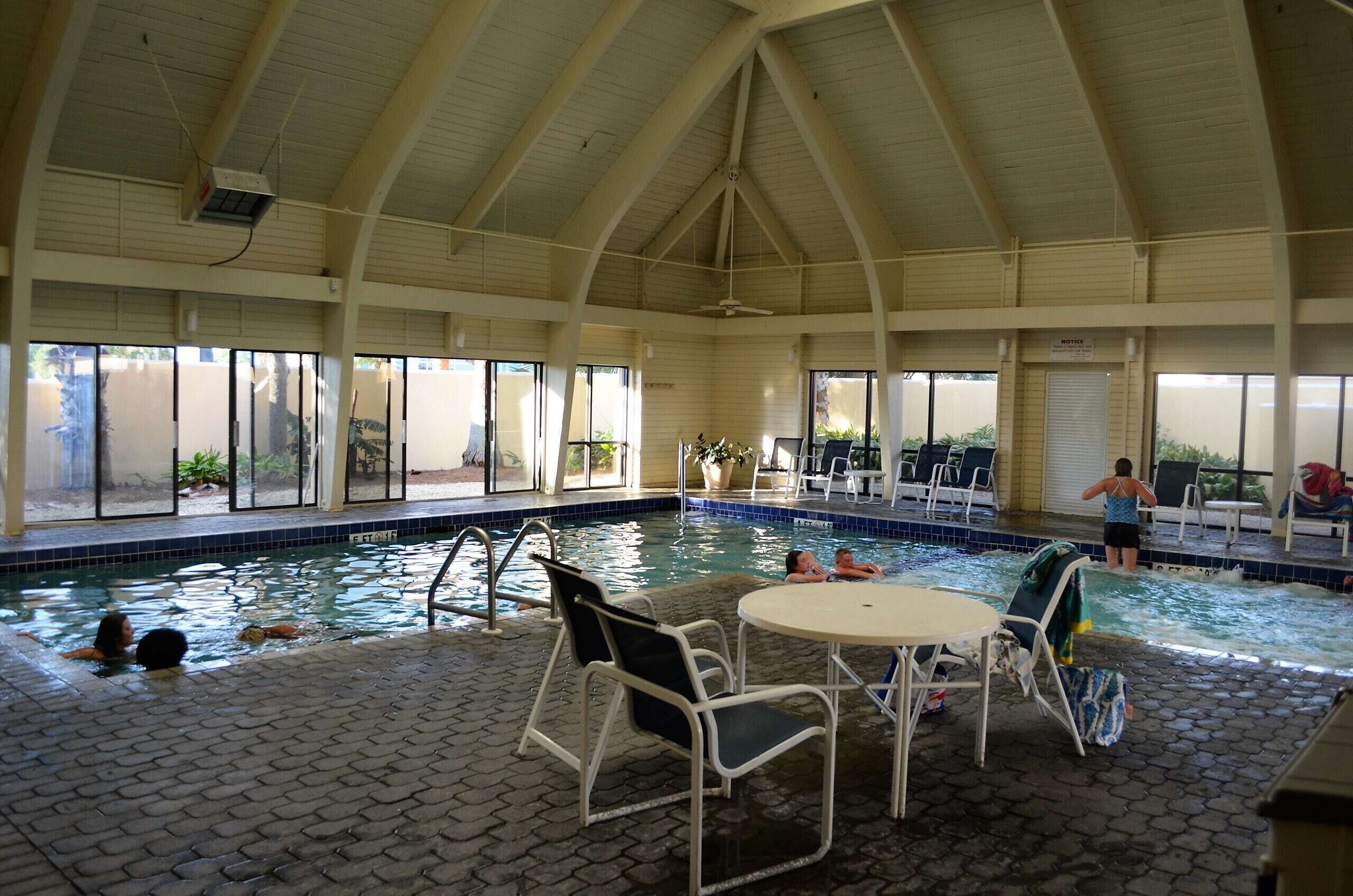 An indoor swimming pool with tiled walls, a vaulted ceiling, and a seating area with chairs and a table.
