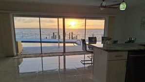 Een ruime kamer met een grote schuifpui naar een balkon met uitzicht op de ondergaande zon boven het water.