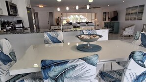 Een moderne eethoek met een witte tafel en stoelen met blauwe bladmotieven, een centrale schaal met fruit, en een keuken op de achtergrond.