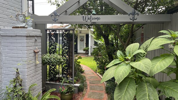 A garden entrance with a wrought iron gate, a brick wall, and a pathway leading to a house.