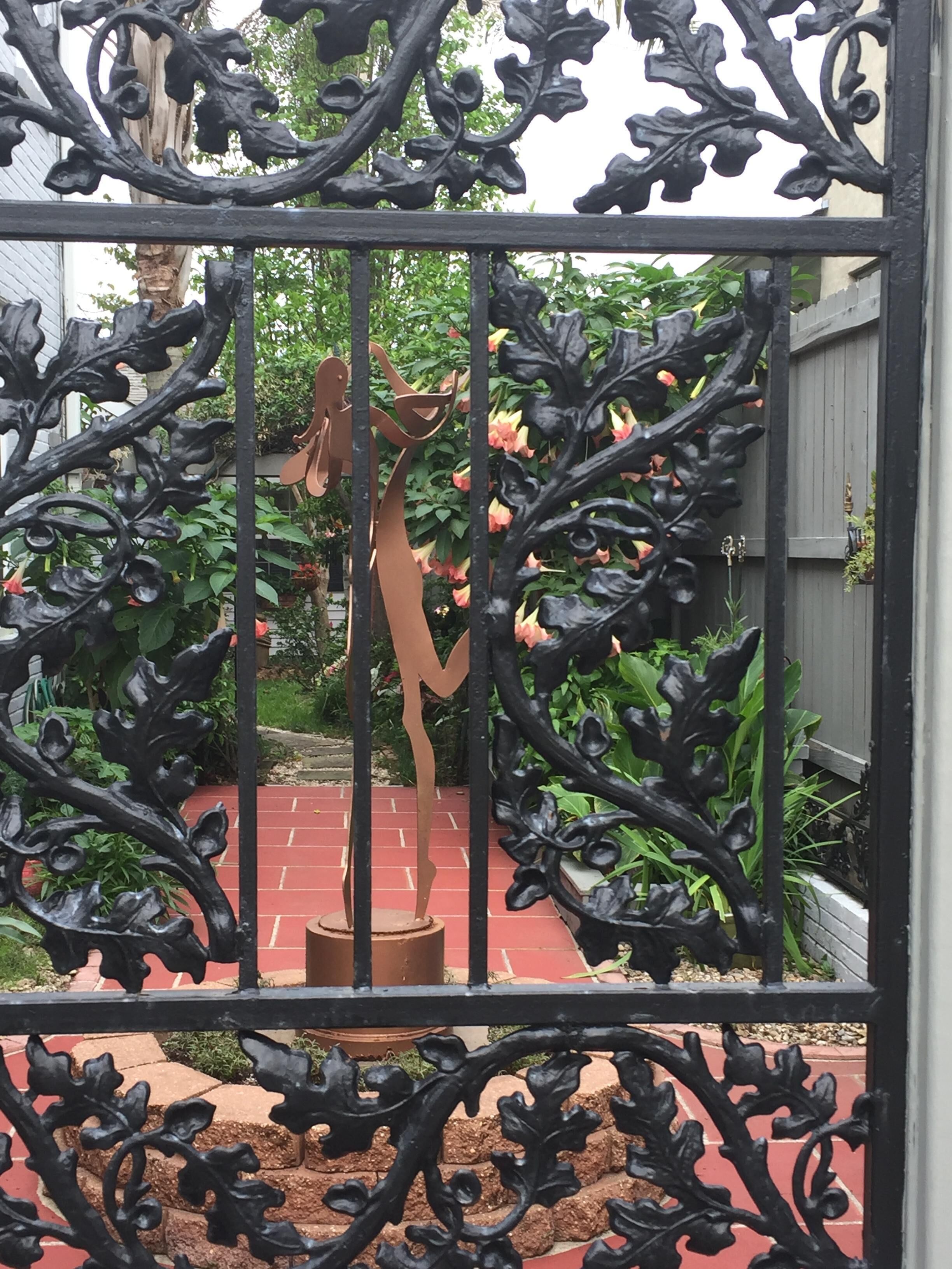 Een ijzeren poort met een decoratief metalen beeld van een figuur met een staf, gelegen in een tuin met rode baksteen vloeren en groene beplanting.