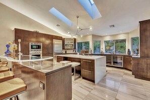 Une cuisine moderne dotée d’un îlot central, d’appareils électroménagers en acier inoxydable et de grandes fenêtres offrant une vue sur la verdure.