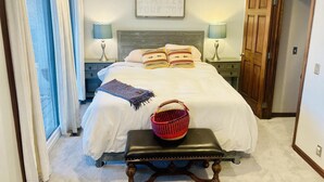 Un dormitorio con una cama, dos mesitas de noche con lámparas, una puerta de madera y una ventana con cortinas.