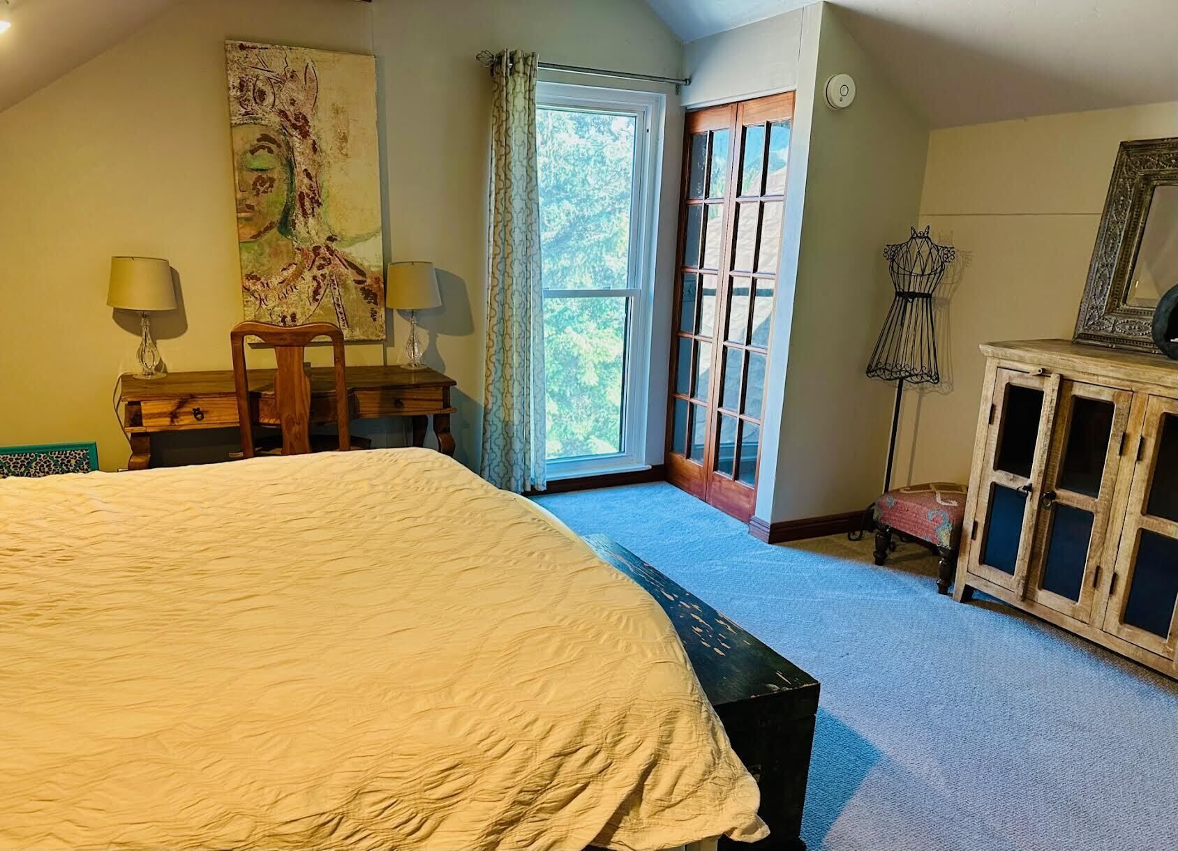 A bedroom with a large bed, a desk with two lamps, a window with curtains, and a cabinet with a mirror.