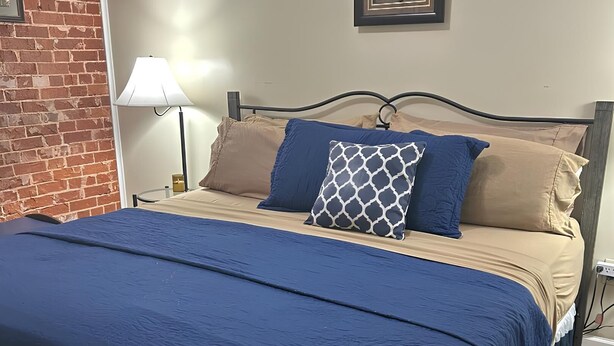 Ein ordentlich bezogenes Bett mit blauer Bettdecke und mehreren Kissen, einer Nachttischlampe und einem gerahmten Bild an der Wand.