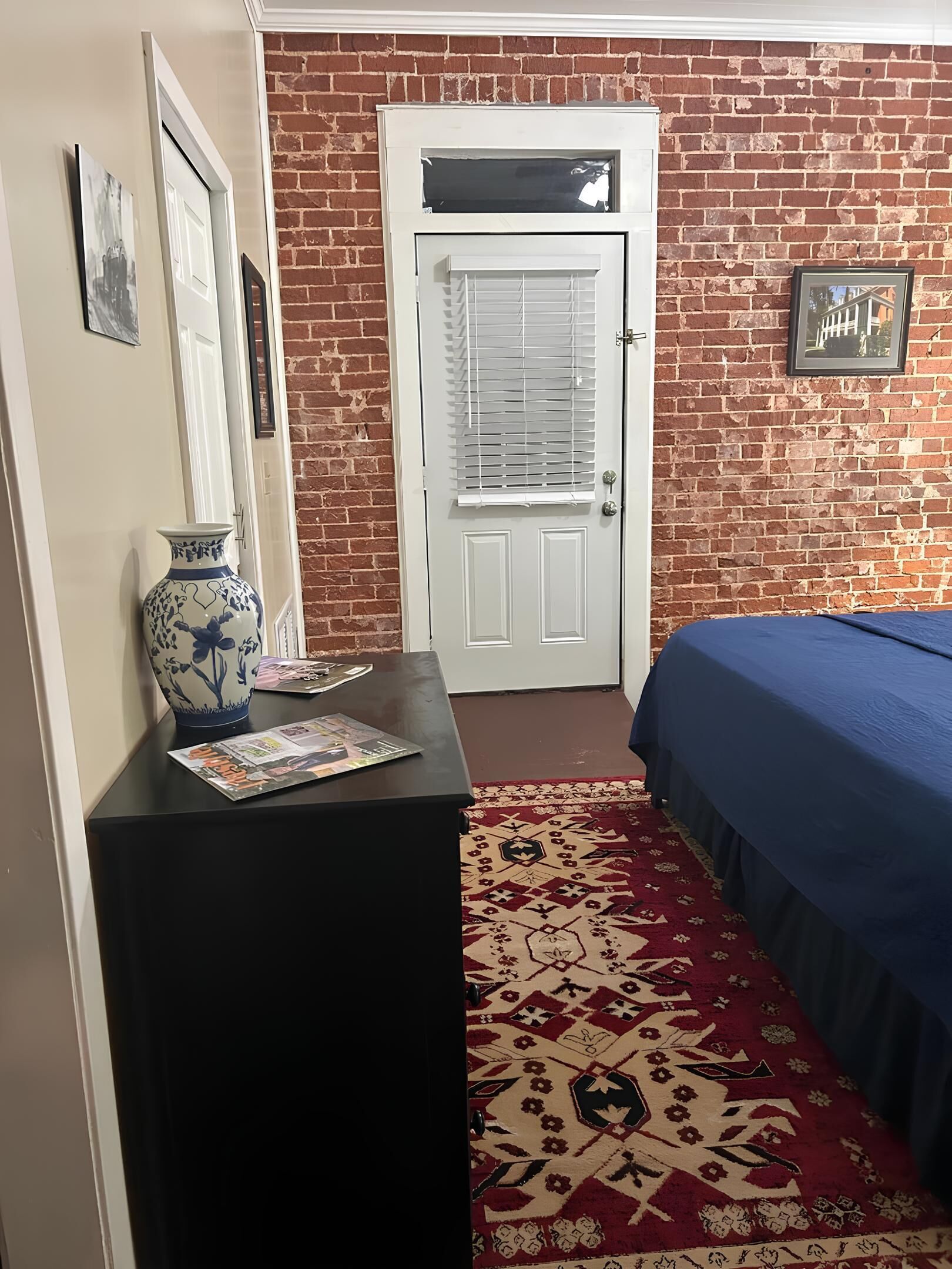 Une pièce avec un mur en briques, une porte blanche, un lit recouvert d’une couverture bleue, une table noire ornée d’un vase et de magazines, et un tapis à motifs.