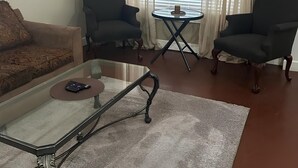 Ein Wohnzimmer mit einem Glastisch, zwei Sesseln, einer Couch, einem kleinen runden Tisch mit Lampe und einem Fenster mit Jalousien.
