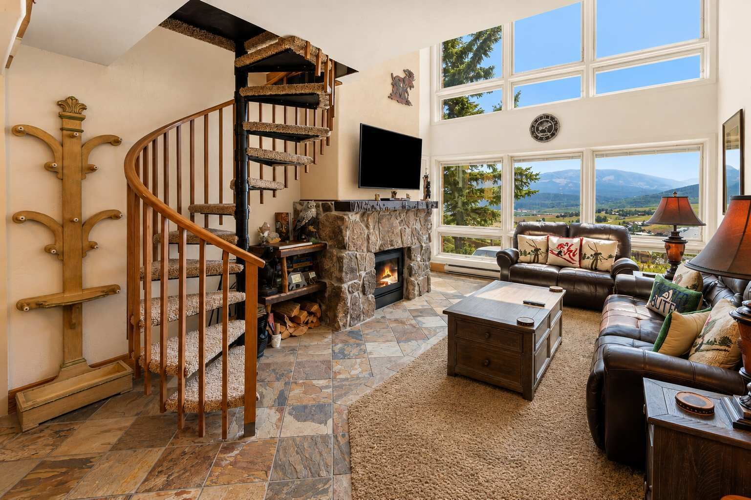 A spacious living room with a stone fireplace, a wooden staircase, a leather sofa, and large windows offering a view of the outdoors.
