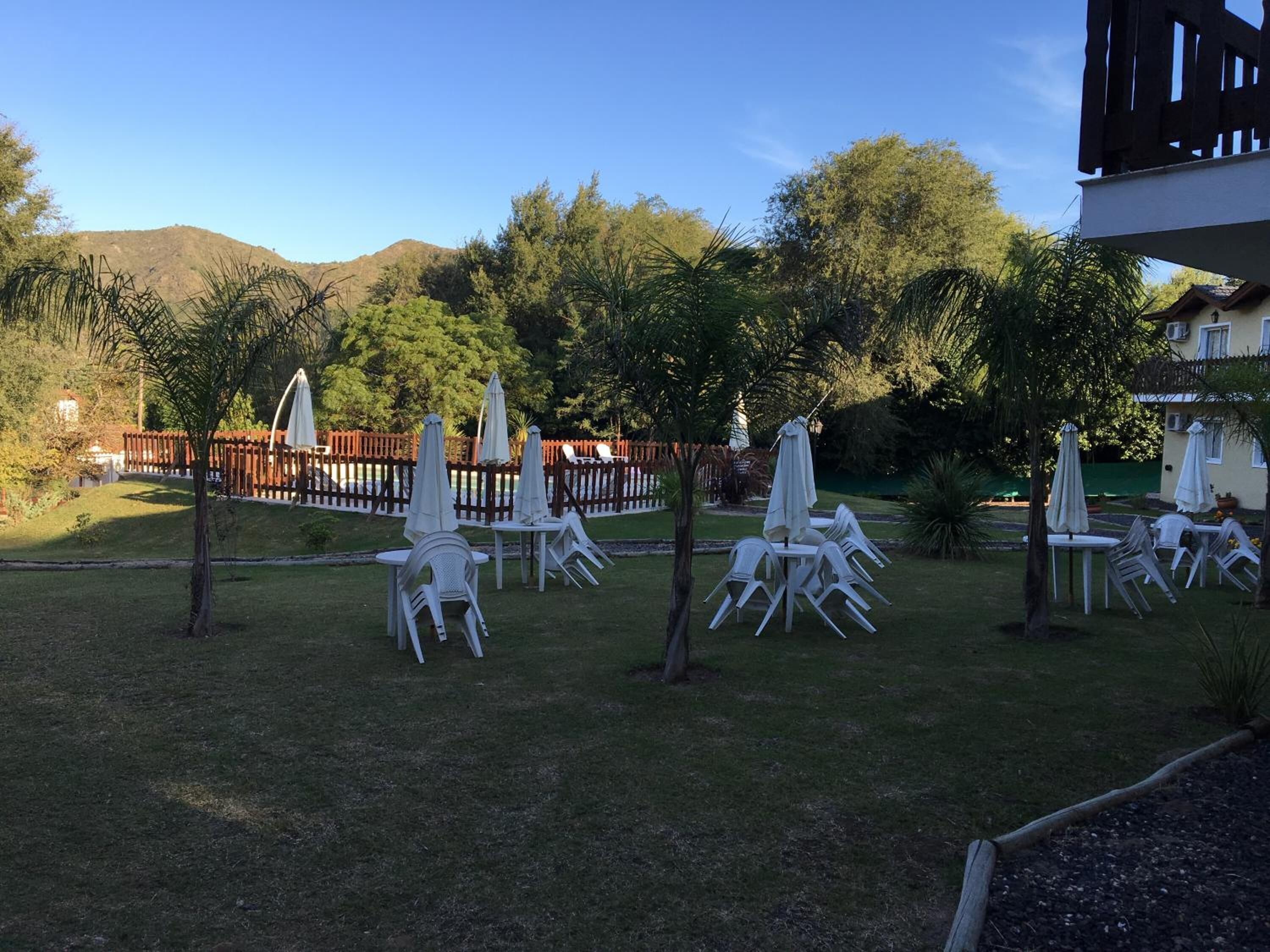 An outdoor seating area with white plastic chairs and umbrellas, surrounded by a wooden fence and trees, with a mountain in the background.
