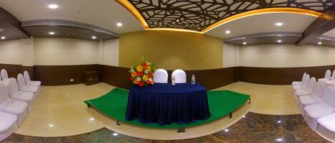 Un salón de conferencias con un escenario, sillas blancas y una mesa con un arreglo floral.