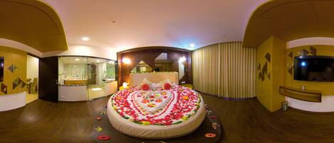 A circular bed with a heart-shaped floral arrangement, a TV, and a bathroom with a glass shower.