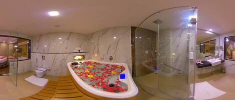 Un baño con pared de mármol, una bañera con pétalos de flores, un box de ducha de vidrio y un lavabo con espejo.