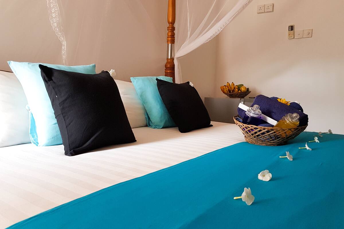 A neatly made bed with a teal bedspread and black and light blue pillows. A wicker basket with toiletries is on the bed.