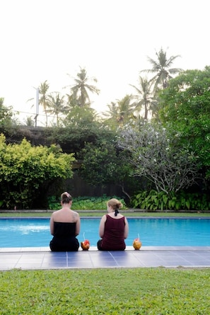 Two people sitting by a pool with drinks, surrounded by lush greenery and palm trees.