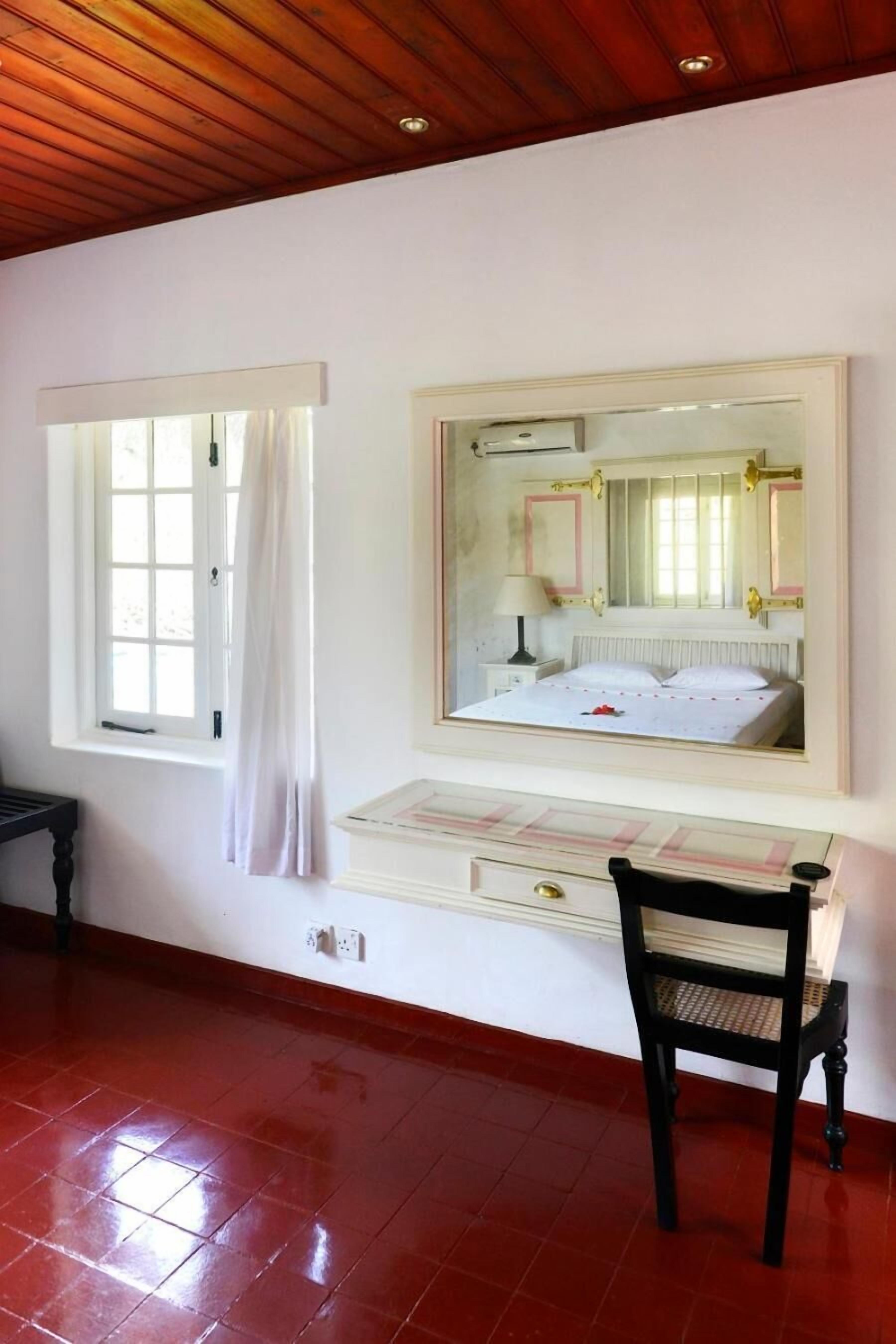 A bedroom with a wooden ceiling, a window with white curtains, a bed with a white duvet, a bedside table with a lamp, and a chair.
