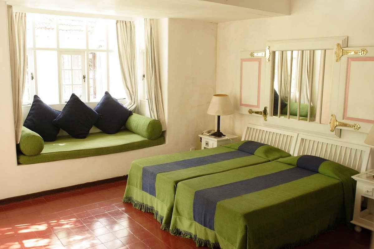 A room with a bed, a window seat with cushions, a bedside table with a lamp, and a radiator.