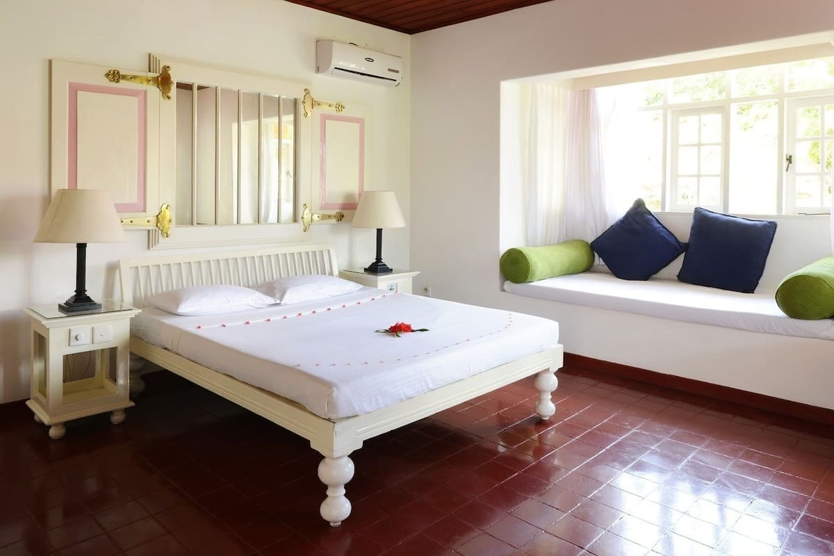 A neatly arranged bedroom with a bed, bedside lamps, a window seat with cushions, and a radiator.