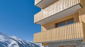 A multi-story wooden building with balconies, stone walls, and a snowy mountain backdrop. - Appart cosy et sympa, à 50m des pistes, sauna (VALMEINIER)