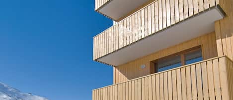 A multi-story wooden building with balconies, stone walls, and a snowy mountain backdrop.