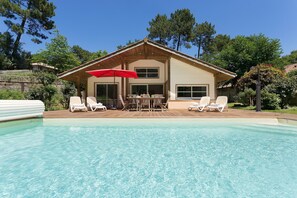 Uma área ao lado da piscina com deck de madeira, um alpendre coberto com um guarda-sol vermelho e uma casa com grandes janelas.