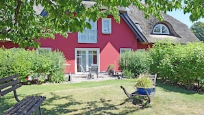 Ett rött hus med ett halmtak, en trädgård med en bänk och en skottkärra full med växter.