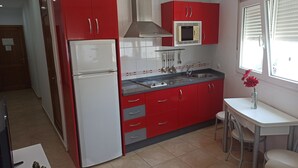 A compact kitchen with red cabinets, a white refrigerator, a microwave, and a sink. There is a small table with a vase of flowers and two chairs. - Terrace with Breathtaking Views - Apartment Sebastian (conil de la frontera)