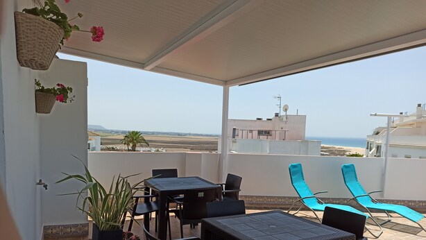 A covered outdoor seating area with tables and chairs, a potted plant, and a view of the sea and distant landscape. - Terrace with Breathtaking Views - Apartment Sebastian (conil de la frontera)