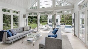 A spacious living room with a grey sofa, a coffee table, and a view of a backyard with a pool and outdoor furniture. - Stunning Hampton Holiday Family Fantasy for your Team  (East Hampton)
