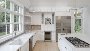 A modern kitchen with white cabinetry, a central island with a stove, and a large window providing natural light. - Stunning Hampton Holiday Family Fantasy for your Team  (East Hampton)