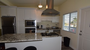 A kitchen with white cabinets, a stainless steel refrigerator, a microwave, a kettle, and a window with blinds. - OCEAN VIEW, 1BR, OUTSTANDING LOCATION F (Carlsbad)