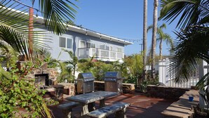 An outdoor patio with stone benches and a grill area, surrounded by palm trees and a building with a balcony. - OCEAN VIEW, 1BR, OUTSTANDING LOCATION F (Carlsbad)