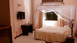 A bedroom with a bed under a canopy, a bedside table with a lamp, a TV mounted on the wall, and a window with curtains. - Golden Peace Hotel (Gulu)