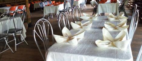 Un salón de banquetes con largas mesas dispuestas para un evento formal, sillas de metal y un interior de madera rústica.